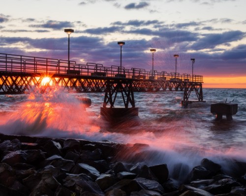 A sunset over a pier, with waves crashing against rocks and vibrant colors in the sky.