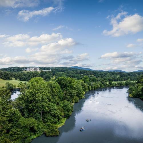 A serene river flows through lush greenery, with a distant view of a grand building under a blue sky with clouds.