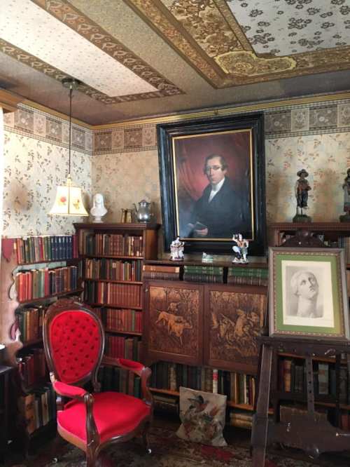 A cozy library with bookshelves, a red chair, a large portrait, and decorative wallpaper.
