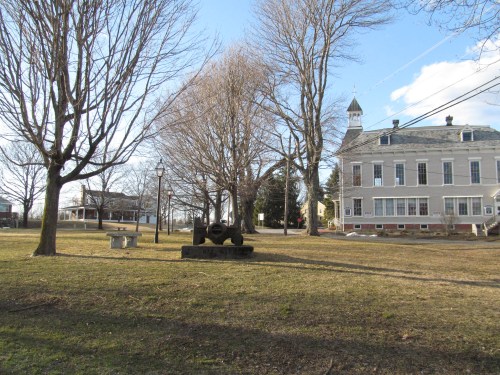 A park scene featuring bare trees, a cannon on a stone base, and a historic building in the background under a clear sky.