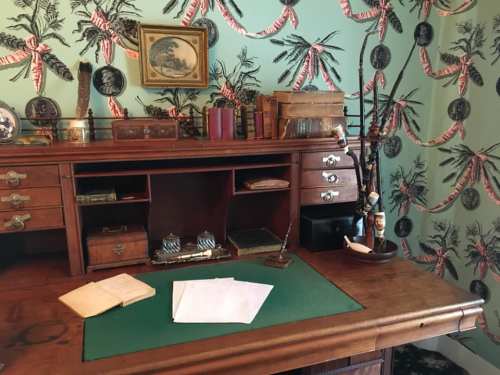 A vintage wooden desk with green blotter, papers, and decorative items, set against a patterned wallpaper.