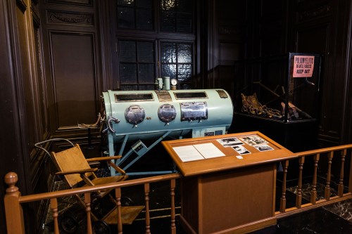 A vintage iron lung machine displayed in a museum, with a wooden table and historical photos nearby.