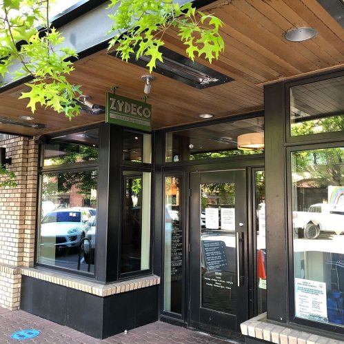 Exterior of a restaurant named "Zydeco," featuring large windows and a wooden ceiling, surrounded by greenery.