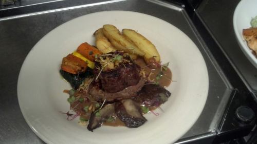 A plated dish featuring a beef patty, roasted vegetables, and crispy potato wedges, garnished with microgreens.