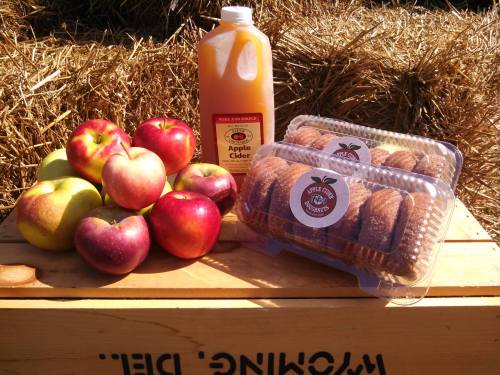 A display of fresh apples, a bottle of apple cider, and a pack of apple cider donuts on a wooden crate.