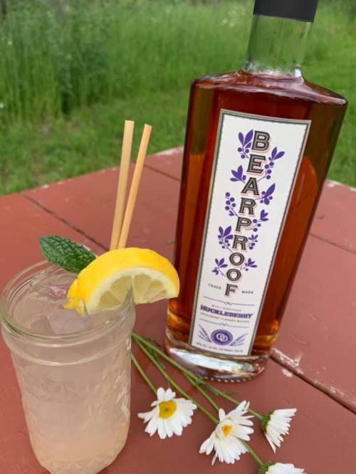 A refreshing cocktail with a lemon slice and mint, next to a bottle of Bearproof Huckleberry spirit and daisies.