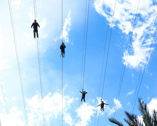 Five people zip-lining across a bright blue sky with scattered clouds, suspended above the ground.