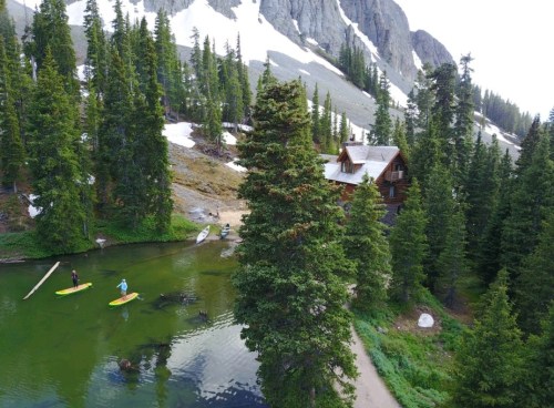 A serene landscape featuring a cabin by a green lake, surrounded by tall trees and snow-capped mountains. Two people paddleboarding.