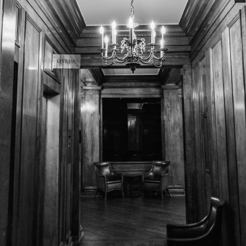 A dimly lit hallway with wooden paneling, featuring a chandelier and two chairs near a door labeled "Gentlemen."
