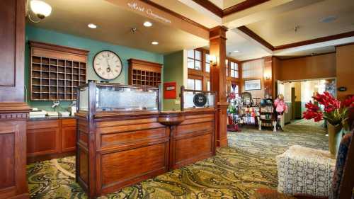 A hotel lobby with a wooden reception desk, clock, and colorful decor, featuring a seating area and gift shop.