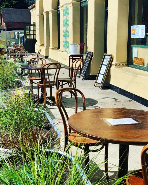 Outdoor seating area with wooden tables and chairs, surrounded by greenery and a building facade in the background.