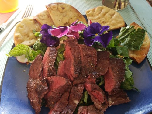 A plate of sliced steak on a bed of greens, garnished with purple flowers and accompanied by flatbreads.