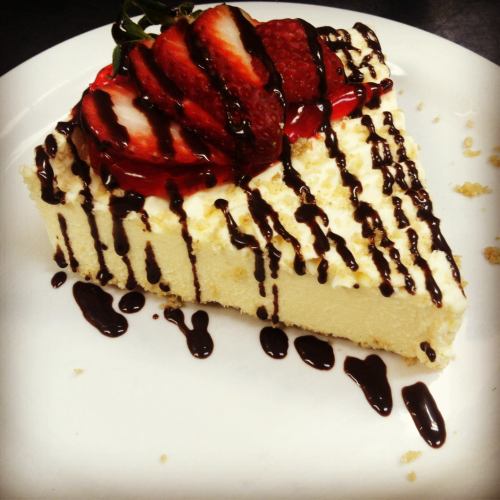 A slice of cheesecake topped with strawberries and drizzled with chocolate sauce on a white plate.
