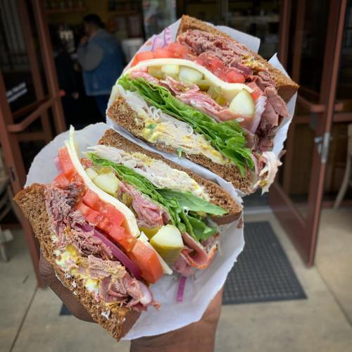 A hand holding a large sandwich filled with layers of meats, lettuce, tomatoes, and pickles, served on dark bread.