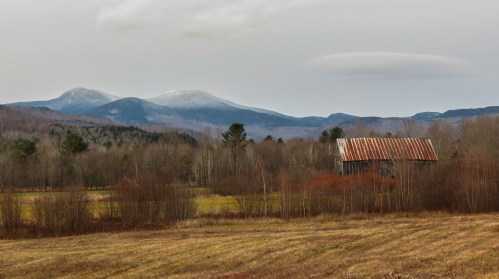 A rustic barn in a field with mountains in the background, partially covered in snow under a cloudy sky.
