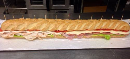 A long sub sandwich with layers of turkey, ham, cheese, lettuce, and tomato, served on a sesame seed hoagie roll.