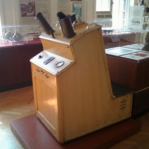 A vintage wooden device with two large tubes, featuring a control panel, displayed in a museum setting.