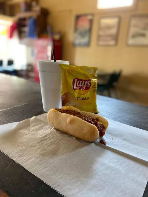A hot dog with chili on a paper napkin, a bag of Lay's chips, and a drink in a cup on a table.