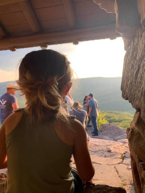 A child sits in a rustic shelter, gazing at a scenic view of mountains and people in the distance during sunset.