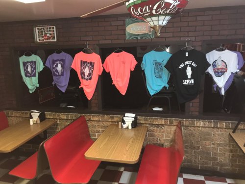 A row of colorful t-shirts hanging in a diner, with red booths and a vintage Coca-Cola sign in the background.