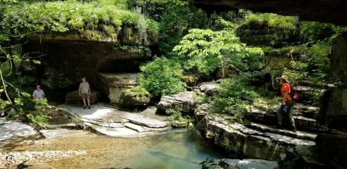 Two people stand near a serene, rocky area with lush greenery and a small stream flowing through.