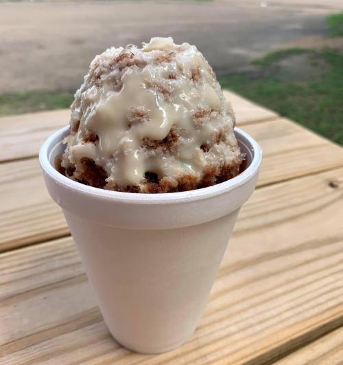A cup of shaved ice topped with a creamy, sweet sauce, sitting on a wooden table outdoors.