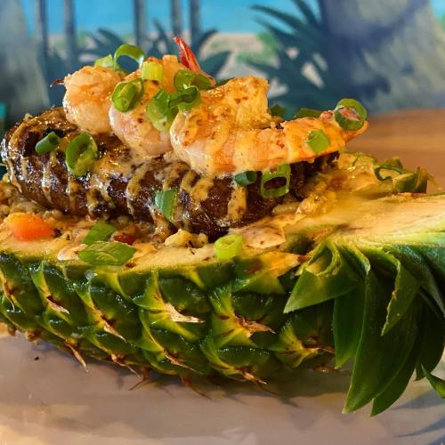 Grilled shrimp and fish served on a halved pineapple, garnished with green onions and a drizzle of sauce.