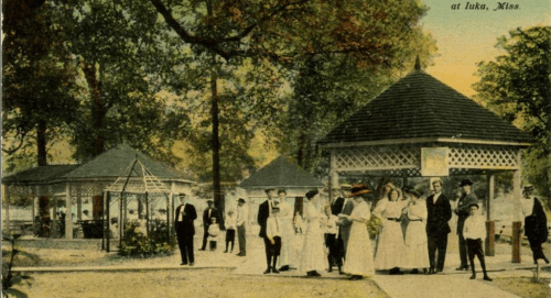 A vintage postcard showing a park scene in Iuka, Mississippi, with people in period clothing near gazebos and trees.
