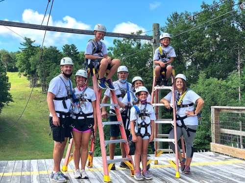 A group of seven people in harnesses and helmets pose on ladders at a zipline course, surrounded by trees and blue sky.