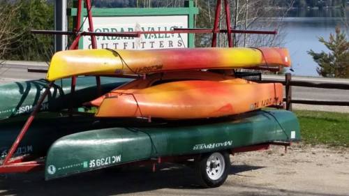 A stack of colorful kayaks on a trailer near a lake, with a sign for "Jordan Valley Outfitters" in the background.