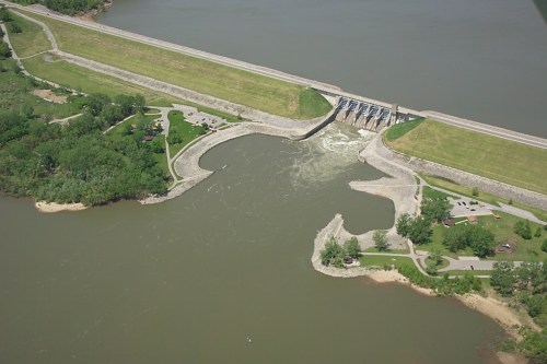 Aerial view of a dam with water flowing through, surrounded by green land and a river.