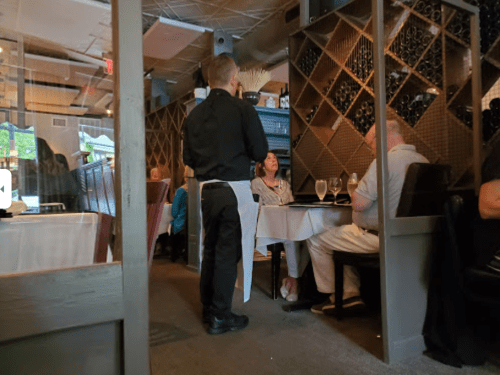 A waiter serves a table in a cozy restaurant with wine racks and diners enjoying their meals.
