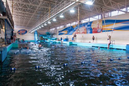 Indoor swimming pool with a slide, swimmers, and lifeguard stations under bright lights and colorful murals.