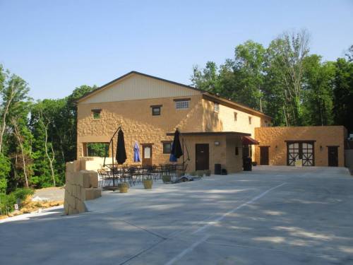 A two-story building with a tan exterior, surrounded by trees, featuring outdoor seating and umbrellas.