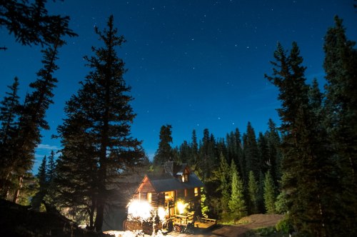 A cozy cabin illuminated at night, surrounded by tall trees and a starry sky.
