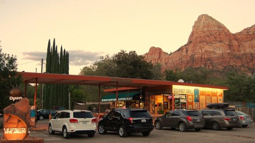 A colorful restaurant with outdoor seating, surrounded by mountains and parked cars at sunset.
