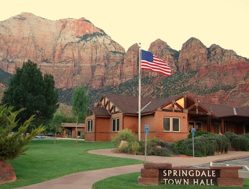 A town hall building in Springdale with an American flag, set against a backdrop of red rock mountains.