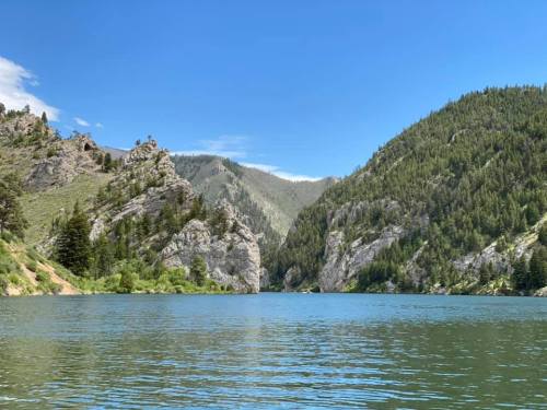 A serene lake surrounded by lush green mountains and rocky cliffs under a clear blue sky.