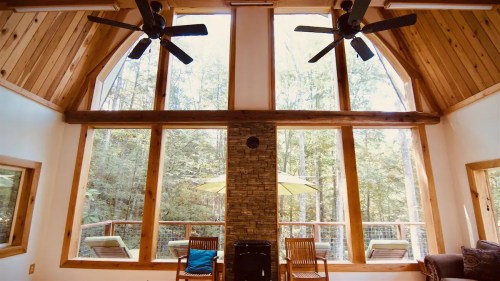 Cozy cabin interior with large windows, wooden beams, and ceiling fans, overlooking a forested deck with lounge chairs.