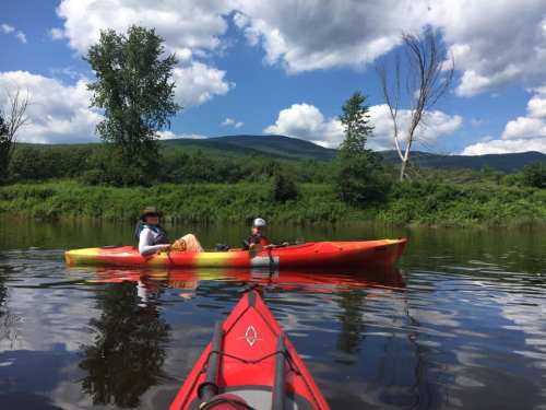 Two people kayaking on a calm river surrounded by lush greenery and mountains under a blue sky with fluffy clouds.