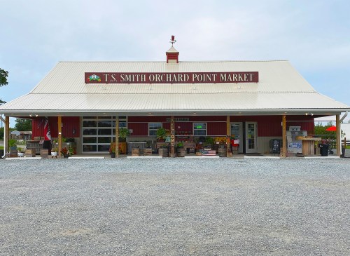 A red barn-style market with a metal roof, featuring a sign that reads "T.S. Smith Orchard Point Market" and gravel parking.