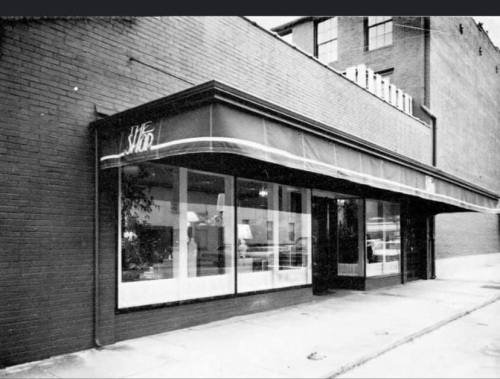 Black and white image of a storefront with large windows and an awning, set against a brick building.