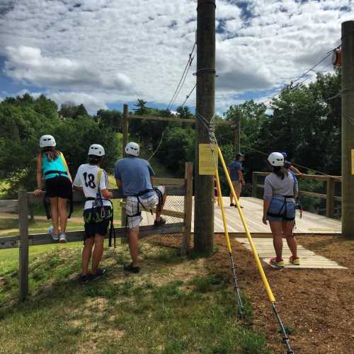 A group of children in helmets and harnesses observe an outdoor ropes course under a cloudy sky.