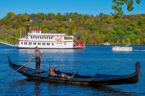 A gondola with passengers on a river, with a steamboat and lush green hills in the background under a clear blue sky.