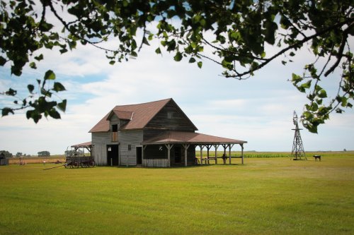 A rustic wooden barn with a porch, surrounded by green fields and a windmill under a blue sky with scattered clouds.
