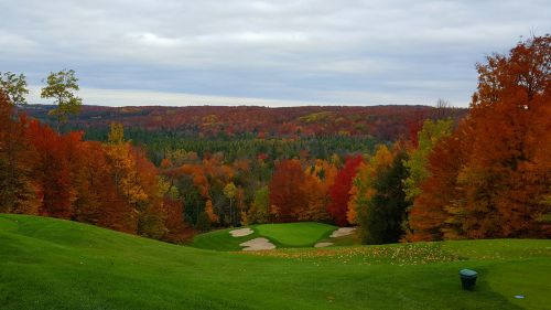A scenic view of a golf course surrounded by vibrant autumn foliage in shades of red, orange, and yellow under a cloudy sky.
