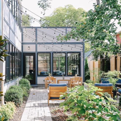 A cozy outdoor patio with string lights, wooden chairs, and greenery, surrounded by a modern building.