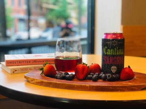 A wooden platter with strawberries, blueberries, a glass of red drink, and a can of Cantina beer, with books in the background.