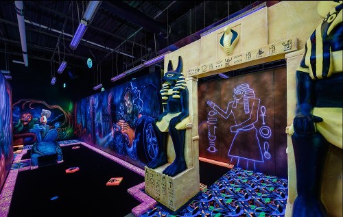 A vibrant indoor mini-golf course with Egyptian-themed murals, statues, and neon lighting.