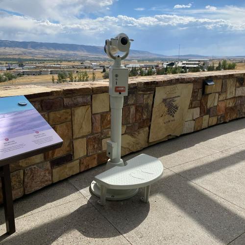 A viewing telescope on a stone platform overlooking a scenic landscape with mountains and blue sky.